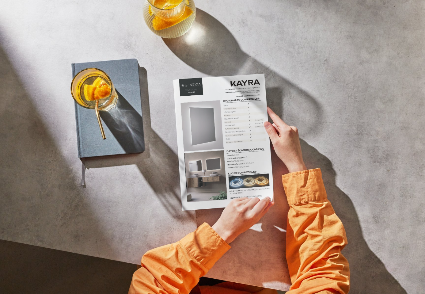 Vista cenital de unas manos sosteniendo la ficha técnica impresa del espejo a medida KAYRA de Genexia sobre una mesa de estilo industrial.